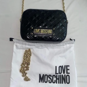 Love Moschino handväska - Säljer denna snygga svarta hand väska från  Moschino. Använd ett fåtal gånger. Har tyvärr inte kvitto kvar för det var en present. Ordinarie pris: 1500 kr. Jag säljer väskan för 1000 kr! Kan mötas upp i Malmö men även frakta men köparen står för frakt. 💕🥰