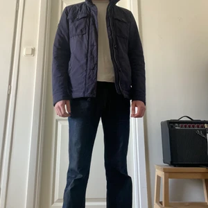 J. Lindberg jacka strl M - Mörkblå jacka från J. Lindberg i storlek Medium, med luva som går att fälla in i kragen. Slitage på ett par ställen, två knappar saknas, inklusive ett hål under högra fickan. Modell: 183 cm