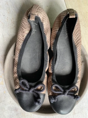 Guldglänsande ballerina stl 38 - Superfin ballerinasko från Clarks. Ormskinnsliknande skinn med guldfärg på nougatfärg. Resårkant runt foten gör att de sitter perfekt och inte glappar. Eleganta till en klänning- läckra till ett par jeans! Inköpta i England- finns inte i Sverige