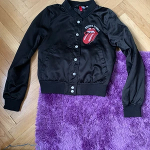 Rolling stones jacka - Superfin Rolling Stones jacka i tunnare glansigt material. Sparsamt använd så väldigt fint skick. Enda defekten är att en av tryckknapparna ramlat av, se sista bilden. Därav det lägre priset. 