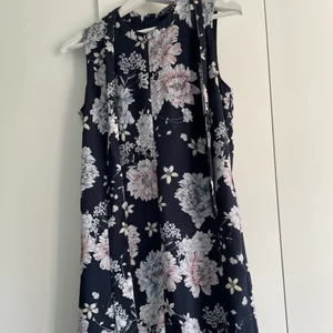 Blommig sommarklänning från Stockholm LM - Fin sommarklänning med stora blommor och mörkblå bas, utan ärmar och med ett medföljande band i samma mönster, dragkedja i nacken. 