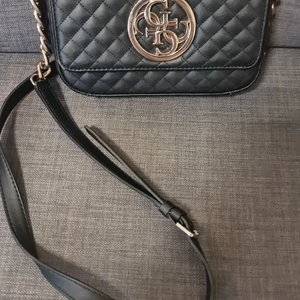 Guess axelremsväska  - Väskan är i bra skick och det finns endast en liten skada på läderkanten och lite missfärgning på två länkar i kedjan . 