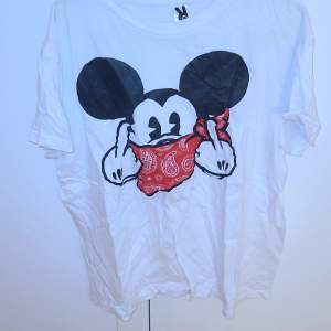 Det är en vit t-shirt men en Mickey Mouse på med röd band runt munnen och sen pekar den med långfigret på båda händerna. 