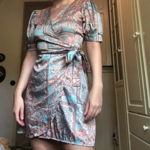 Omlottklänning - Fin omlottklänning i silkesmaterial. Den har knytning i midjan och puffärm, har även satt en säkerhetsnål vid urringningen. Storlek 40/L. 