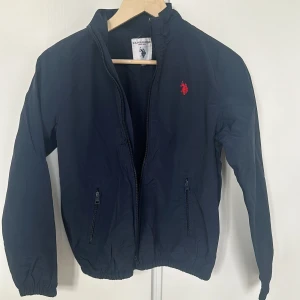 Mörkblå jacka från U.S. Polo Assn. - Säljer en snygg mörkblå jacka från U.S. Polo Assn. med dragkedja och två fickor framtill. Jackan har en röd broderad logga på bröstet och är perfekt för vår och höst. Den är i bra skick och passar perfekt för en casual look. Storleken är 12-13 YRS