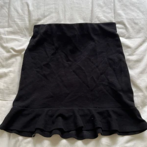 Svart kjol med volang - Säljer en snygg svart kjol med volangdetalj längst ner. Kjolen är tight och passar perfekt för både vardag och fest. Den är i bra skick och har en enkel men stilren design. Storlek 158/164 i barn men passar mig som har XS/S