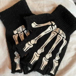 Svarta fingerlösa handskar med skelettmönster - Säljer ett par coola svarta fingerlösa handskar med ett vitt skelettmönster på ovansidan. De har en nätliknande design på handflatan som ger en edgy look. Perfekta för att ge din outfit en unik touch!