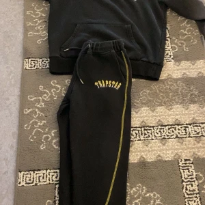 Svart hoodie från Trapstar - Säljer en svart hoodie från Trapstar med gul text på bröstet. Den har en klassisk känguruficka och ribbade muddar vid ärmslut och nederkant. Perfekt för en chill dag eller när du vill se stilren ut. Passar bra till både vardag och fest!