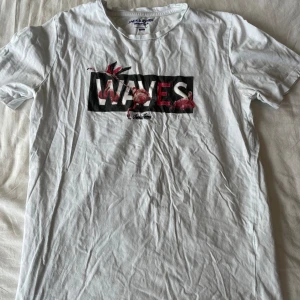 Vit t-shirt från Jack & Jones - Säljer en vit t-shirt från Jack & Jones med trycket 'WAVES' och några coola flamingos på framsidan. Den är i storlek L och har korta ärmar. Perfekt för sommaren eller en avslappnad dag. T-shirten är i bomull och har en normal passform. Den är lite skrynklig men annars i bra skick. Passa på att köpa denna stiliga t-shirt! (Passar M) Frågor? Skriv!