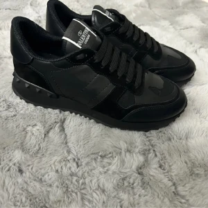 Svarta sneakers från Valentino Garavani - Säljer ett par svarta sneakers från Valentino Garavani. Skorna är i nyskick och har en cool design med svarta detaljer och snörning. De är tillverkade i skinn och mocka, vilket ger dem en lyxig känsla. Perfekta för både vardag och fest!