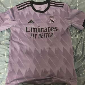 Säljer en snygg lila Real Madrid fotbollströja från Adidas. Tröjan har ett coolt geometriskt mönster och är kortärmad. Den är i nyskick och har 'Emirates Fly Better' tryckt på framsidan. Perfekt för alla fotbollsfans!