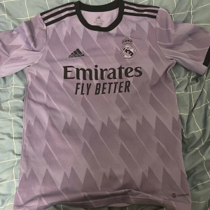 Lila Real Madrid fotbollströja från Adidas - Säljer en snygg lila Real Madrid fotbollströja från Adidas. Tröjan har ett coolt geometriskt mönster och är kortärmad. Den är i nyskick och har 'Emirates Fly Better' tryckt på framsidan. Perfekt för alla fotbollsfans!