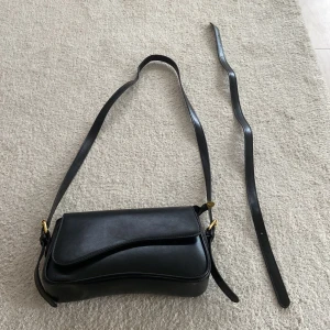 Svart axelväska i skinn - Säljer en snygg svart axelväska i skinnimatiskon . Har aldrig använt.Väskan har en avtagbar axelrem och en dragkedja på insidan för extra säkerhet. Perfekt för både vardag och fest! 