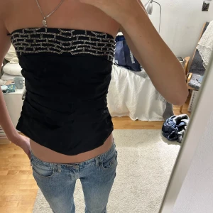 Svart tubtopp med glittriga detaljer - Säljer en snygg svart bandeutopp med pärlor/glittriga detaljer upptill. Toppen är axelbandslös och har en dragkedja på baksidan. Perfekt för festliga tillfällen! Passar bra till jeans eller kjol. 🖤✨