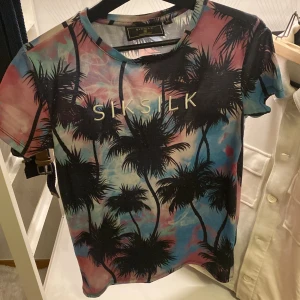 Flerfärgad t-shirt med palmmönster från SikSilk - Säljer en supercool t-shirt från SikSilk med ett tropiskt palmmönster i flera färger som rosa, blå och svart. Perfekt för sommaren med sin kortärmade design och mjuka bomullsmaterial. Den har en normal passform och är i nyskick. Perfekt för en avslappnad dag eller en festkväll!