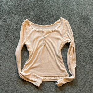 Beige ribbad långärmad topp - Säljer en krämvit ribbad långärmad topp med knappar framtill. Toppen har en rund halsringning och en diskret mönstrad kant på insidan. Perfekt för en avslappnad stil.
