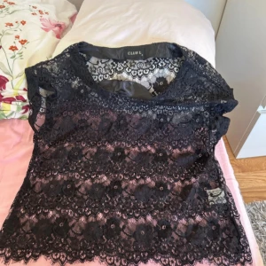 Svart spetsblus från Club L - Elegant svart spetsblus från Club L med korta ärmar och ett vackert blommönster. Perfekt för en stilfull look med en feminin touch. Blusen har en rund halsringning och är tillverkad i ett lätt och luftigt material.