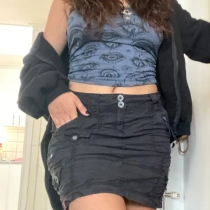 Svart kortkjol med knappar - Snygg svart cargo-kjol med knappar framtill och fickor på sidorna. Perfekt för en cool och avslappnad stil. Kjolen har en enkel design som gör den lätt att matcha med olika toppar.✨