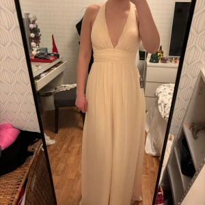 Champagnefärgad Balklänning från NLY Eve - Super fin klänning som passar perfekt till balen. Helt ny med prislappen kvar. Elegant champagnefärgad långklänning från NLY Eve med djup v-ringning där banden korsas i ryggen.