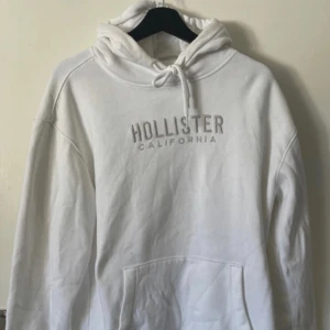 Vit hoodie från Hollister - Säljer en vit hoodie från Hollister med texten 'Hollister California' på bröstet. Den har en justerbar huva med dragsko och en stor magficka framtill. Perfekt för en avslappnad stil.