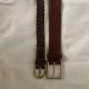 Två bruna bälten i skinn - Säljer två snygga bruna bälten i skinn. Ett är flätat med en rundad guldfärgad spänne och det andra är slätt med ett fyrkantigt silverfärgat spänne. Perfekta för att komplettera din outfit! 95 cm och 100 cm 