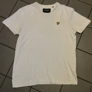 Vit t-shirt från Lyle & Scott - Säljer en klassisk vit t-shirt från Lyle & Scott med korta ärmar och en broderad gul logotyp på bröstet. Perfekt för en stilren look. Liten fläck vid logotype