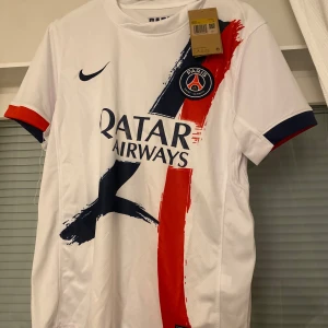 Vit PSG fotbollströja - Snygg vit PSG fotbollströja från Nike med korta ärmar. Tröjan har ett coolt mönster med röda och blå penseldrag och PSG-loggan på bröstet. Perfekt för alla fotbollsfans!