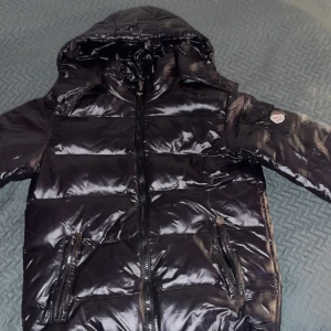 Svart dunjacka från Moncler - Snygg svart dunjacka från Moncler med glansig finish och huva. Jackan har dragkedja framtill och praktiska fickor med dragkedjor. Perfekt för kyliga dagar.