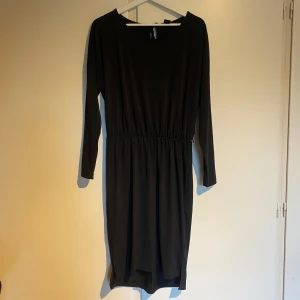 Svart klänning från Lindex - Elegant svart klänning från Lindex med lång ärm och knytband i midjan. Perfekt för en stilren look. Klänningen har en öppning i ryggen och knytning i ryggen.