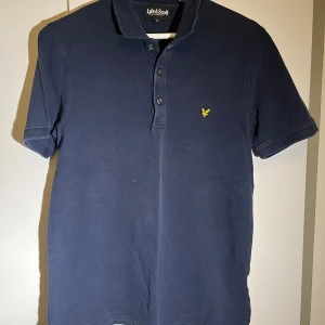 Mörkblå pikétröja från Lyle & Scott - Snygg mörkblå pikétröja från Lyle & Scott med klassisk krage och knappslå. Tröjan har en liten gul logga på bröstet och korta ärmar. Perfekt för en stilren look.