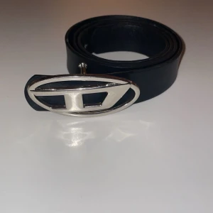 Svart bälte Diesel - Snyggt svart Diesel bälte med ett unikt silverfärgat spänne i metall. Perfekt för att ge din outfit en stilren touch. Bältet har flera justerbara hål för bästa passform.