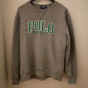 Ralph Lauren - Säljer en grå sweatshirt från Polo Ralph Lauren med grönt tryck på bröstet och Ralph Lauren märket på höger ärm.                                           Storlek 14-16 tröjan är i ny skick.