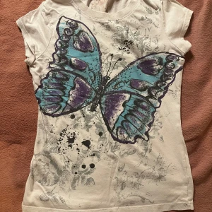 Vit t-shirt med fjärilstryck - Snygg vit t-shirt med ett stort fjärilstryck i blått och lila på framsidan. T-shirten har korta ärmar och en rund halsringning. Perfekt för en avslappnad stil med en touch av färg.