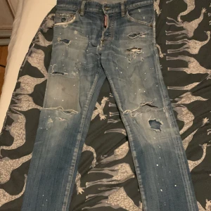 Blå jeans från Dsquared2 - Säljer ett par coola blå jeans från Dsquared2 med slitna detaljer och färgstänk. De har en klassisk femficksdesign och knappgylf. Perfekta för en avslappnad stil med en edgy touch.