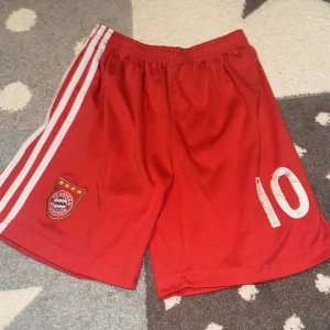Röda fotbollsshorts från Bayern München - Snygga röda fotbollsshorts från Bayern München med vita ränder på sidorna och klubbens emblem på ena benet. Nummer 10 tryckt på framsidan. Perfekta för fotbollsträningen eller matchen!