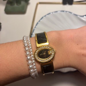 Elegant guldfärgad klocka med svart armband - Snygg guldfärgad klocka med svart armband och glittrande stenar runt urtavlan. Perfekt accessoar för att lyfta din stil. Urtavlan är svart med guldfärgade detaljer. Fungerar!