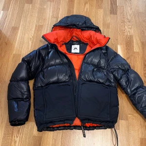 Svart dunjacka från J.Lindeberg - Snygg svart dunjacka från J.Lindeberg med en iögonfallande orange insida. Jackan har en praktisk huva och dragkedja framtill. Perfekt för kyliga dagar. Nypris 5499kr.