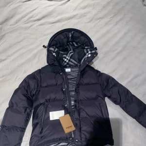 Svart Burberry Puffer Jacka/Väst - Snygg svart dunjacka från Burberry med quiltad design och dragkedja. Jackan har en avtagbar huva med ett klassiskt rutigt mönster i grått och vitt. Perfekt för kyliga dagar. Det är storlek M men är lite liten i storlek. Kan också göras om enkelt till en väst.