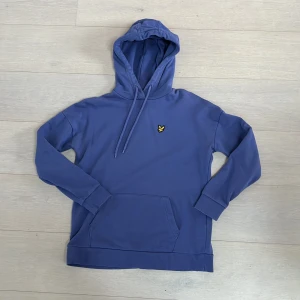 Blå hoodie från Lyle & Scott - Säljer en snygg blå/lila ton hoodie från Lyle & Scott i 100% ekologisk bomull. Tröjan har en klassisk design med en känguruficka och en broderad logotyp på bröstet. Perfekt för en avslappnad stil.