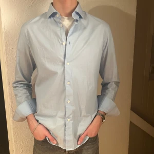 Ljusblå skjorta - Snygg ljusblå skjorta med klassisk krage och knappar framtill. Perfekt för en stilren look. Skjortan har långa ärmar och är tillverkad i ett lätt material. Storlek M, 40. 