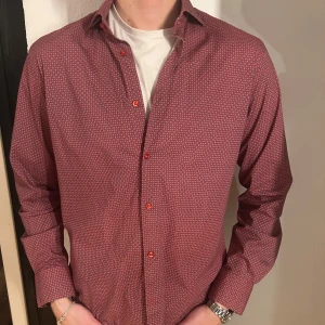 Röd mönstrad skjorta - Snygg röd skjorta med diskret mönster och långa ärmar. Perfekt för en stilren look. Skjortan har knappar framtill och en klassisk krage. Storlek L, 42. Men passar även M 