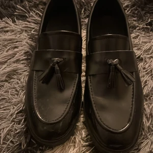Svarta loafers med tofsar -  svarta loafers i skinn med dekorativa tofsar på ovansidan. Perfekta för en stilren look.