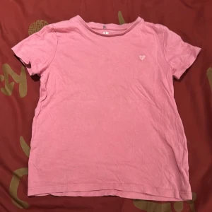 Rosa t-shirt från H&M - Säljer en rosa t-shirt från H&M i ekologisk bomull. Den har en liten hjärtapplikation på bröstet och korta ärmar. Perfekt för vardagsbruk! Katt finns i hemmet. 