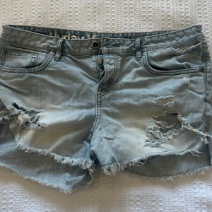 Ljusa jeansshorts från Hurley - Säljer ett par ljusa jeansshorts från Hurley med slitna detaljer och fransiga kanter. Perfekta för en avslappnad stil. Shortsen har klassisk femficksdesign och knappgylf.