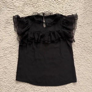 Svart blus med spetsdetaljer - Säljer denna superfina blus med volang och spetsdetaljer. Blusen har med prickigt mönster vid brösten och har en knapp vid nacken. Passar perfekt inför middagar eller en tur på stan!