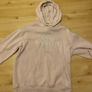 Ljusrosa hoodie från H&M - Säljer en ljusrosa hoodie från H&M med texten 'New York City' broderad på framsidan. Den har en stor ficka framtill och en bekväm huva med dragsko. Perfekt för en avslappnad stil.