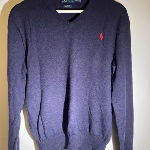 Mörkblå tröja från Ralph Lauren - Säljer en stilren mörkblå tröja från Ralph Lauren i merinoull. Tröjan har en v-ringad design och ett rött broderat märke på bröstet. Perfekt för en klassisk look. Storlek L för barn passar S 
