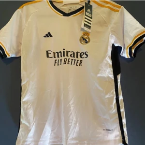 Vit Real Madrid fotbollströja - Säljer en vit Real Madrid fotbollströja från Adidas med korta ärmar. Tröjan har svarta och gula detaljer samt klubbens emblem på bröstet. Perfekt för fans som vill visa sitt stöd. Shorts medföljer 