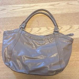 Beige axelväska i syntetmaterial - Snygg beige axelväska i syntetmaterial med dubbla handtag och dragkedja. Perfekt för att bära med sig det nödvändigaste. Stilren design som passar till många tillfällen.