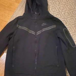 Svart hoodie från Nike - Säljer en svart hoodie från Nike med dragkedja och huva. Jackan har långa ärmar och en stilren design med diskreta detaljer. Perfekt för en sportig look eller avslappnade dagar.